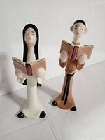 Front view of both vintage ceramic figurines of a singing monk and nun, holding hymn books, standing side by side.