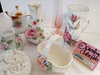 Overall view of the lot showing the porcelain vases, lidded dish shaped like roses, pink glass bud vase, and floral decorated pieces including gold gilt accents.