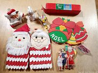 Overview photo of lot showing Santa and Mrs. Claus fabric pot holders, ceramic candleholder, soap dispenser, boxed chimes, fabric Noel wreath and bell, wooden Noel cutouts and other decorations.