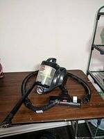 Black Bissell bagless vacuum cleaner with hose and floor brush attachment on brown surface, light background. Clear dustbin visible.