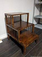 Front and side angled view of wooden 3-tier stepped end table showing three square platforms with slatted vertical bars on sides and medium brown finish.