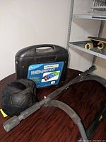 Photo showing the black weighted ball with handles, black plastic case labeled 'Fitness Gear' containing dumbbells, and a black pull-up bar with padded grips all arranged on a brown table with shelving in background.