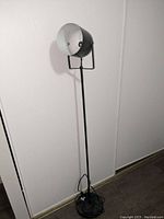 Black metal floor lamp approximately 58 inches tall with adjustable spotlight head and round open base shown against neutral wall.