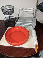 Kitchen items showing marble board, rolling pin, orange bowl, black metal two-tier fruit basket, and white metal two-tier dish rack on wooden table