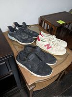 View of three pairs of men's shoes on a table including black Adidas shoes, white New Balance sneakers with red 'N' logo, and black shoes in front.