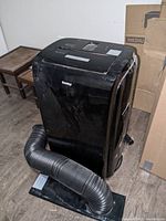 Black Danby portable air conditioner unit with attached flexible black vent hose and rectangular vent base attachment on floor, positioned near boxes and brown side tables.