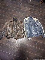 Two vintage men's leather motorcycle jackets laid on dark wood floor, showing brown and blue jackets side-by-side