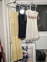 Three women's dresses hanging on hangers: yellow floral maxi, black fitted sleeveless, and white strapless with brown beaded detail.