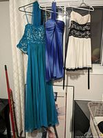 Three women's dresses hanging on hangers: teal long dress with lace upper bodice, blue strapless medium dress, white/cream strapless medium dress with black lace band and trim.