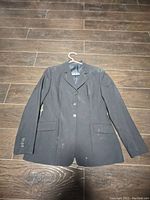 Front view of black women's horseback riding jacket laid flat on wood floor laid on hanger. Shows three buttons down front, two pockets with flaps, some dust and signs of light use.