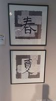 Two framed art prints stacked vertically on a wall, each with Asian calligraphy characters and textured backgrounds.