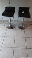 Pair of black seat swivel bar stools with chrome metal base and footrest, shown from front angle on tiled floor.