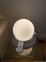 Front view of the electric orb lamp on its silver textured stand on a glass table beside a beige couch.