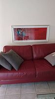 Framed abstract painting hanging above a red leather couch with pillows.