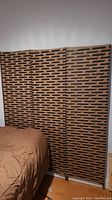 Full view of three panel woven rattan room divider placed upright beside a bed and wall.