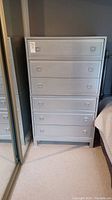Front view of the silver 6 drawer tall boy dresser placed near a mirrored closet and bed.