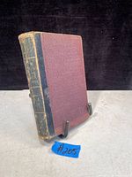 Front cover and spine of the antique book showing maroon cloth cover with black leather spine with wear.