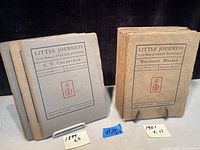 Three 1899 volumes of Little Journeys books with gray covers on left and eleven 1901 volumes with tan covers on right, displayed upright on stands