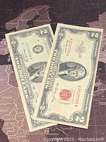 Two US $2 banknotes lying on a map, displaying front side with portraits and seals.