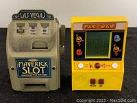 Two small vintage handheld gaming systems shown side by side on white surface. Left is a Maverick Slot small slot machine game with lever and Las Vegas sign. Right is a yellow Pac-Man handheld electronic game with maze display and character graphics.