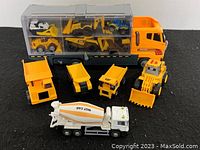 Front view of the large orange and gray carrier truck with its transparent multi-compartment trailer holding small construction vehicles mainly yellow CAT toys, and white cement mixer truck and several larger machines positioned in front.