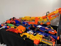 Overview of numerous Nerf blasters piled on a black surface, showing different styles and colors including orange, yellow, blue, red.