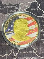 Front side of gold coloured medallion showing Donald J. Trump's profile with American flag background and inscriptions.