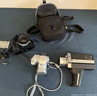 Three vintage cameras on black background: Bell & Howell camcorder, Canon camera, black SLR camera with carrying case.