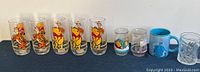 Photo shows a row of collectible glasses featuring Tigger, Winnie the Pooh, a Niagara Falls stein, a Cinderella ceramic mug, and other character-themed glasses.
