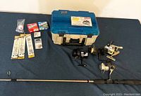 Full display of the fishing tackle box, three fishing reels, fishing rod, fishing line boxes, hooks, leaders, and bobber arranged on a dark surface.