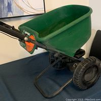 Side view of Scott's basic broadcast spreader showing green plastic hopper, black frame with wheels, handlebar, and orange adjustment dial