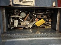 Open metal toolbox showing a jumble collection of assorted hand tools and hardware including a coil of yellow string, bolts, clamps, and a hammer head.