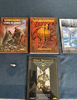 Four Warhammer books displayed on blue surface: 'Storm of Chaos', book with helmet on cover, 'Index Astartes IV', and 'Imperial Armour' booklet. Also includes small metal figure base visible at edge.