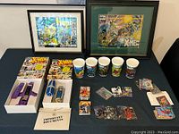Overview of entire lot showing framed comic covers, collectible cups, two watches in boxes, trading card packs, and loose cards arranged on a table.