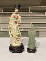 Two Asian resin figurines, one 7 inches tall woman in traditional attire on wooden base, and one 4 inches tall translucent green robed man on dark base.