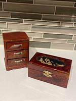 Photo showing the two Asian lacquer boxes side by side. The rectangular box has a cork carving on the lid and clasp, the chest has 3 drawers with metal handles.