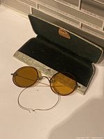 Antique round sunglasses with amber lenses displayed inside open hard case showing the interior lining and metal clasp.