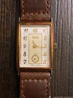 Close-up photo of the Gruen Precision rectangular watch face showing gold-tone case, white dial with gold Arabic numerals, and brown leather strap.