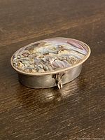 Oval pill box viewed at an angle showing the textured iridescent shell inlay on the lid and metal body with clasp.