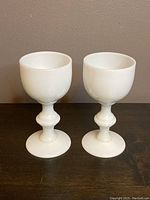 Front view of two identical mini milk glass shot glasses, white opaque with goblet style and decorative stems, standing side by side on a dark surface against a beige wall.
