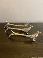 Photo showing three silver plated pheasant-shaped utensil rests positioned side by side on a dark wooden surface, highlighting detailed feather design and elongated shape.