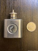 Front view of tiny stainless steel fishing flask with embossed fish image and a Canadian quarter for size reference indicating 1oz capacity.