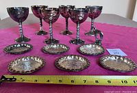 Photo of six silver-plated wine goblets arranged in two rows, front row shows detailed twisted stems and rounded bowls, placed on a pink fabric with measuring tape showing length just over 10 inches across the group.