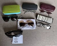 Seven pairs of glasses shown with four cases on a soft surface, includes a mix of sunglasses, reading and prescription glasses.
