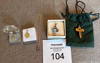 Four pendants presented on fabric background: small gold cross pendant in clear plastic box, gold infant Jesus pendant on chain on white cushion, colorful painted glass cross pendant in a box, and wooden cross pendant on black cord pouch.