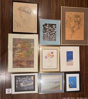 Nine picture frames laid out on floor showing mix of artwork: pencil sketches, prints, and paintings. Frames vary in color and condition with some signs of damage and wear.