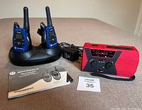 Photo showing entire lot including red and black emergency weather radio, two blue Motorola walkie-talkies in a charging dock, power adapter, and instruction manual on a brown surface.