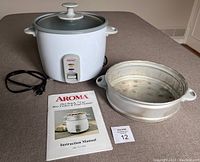 Rice cooker, steamer insert, and instruction manual displayed separately on table.