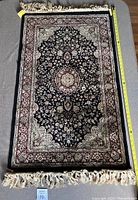 Front side of rectangular viscose pile rug with dark central pattern and floral border, approx. 65 by 110 cm with fringe on ends. Measuring tape placed for scale.