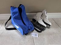 Pair of white Lange Galaxy women's ice skates with blades showing rust, placed next to blue Bauer skate bag on tiled floor.